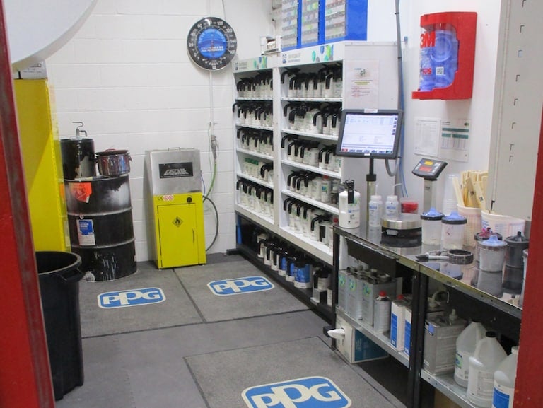 A well-stocked auto body paint mixing room with shelves of paint, industrial equipment, and PPG logos on the floor.